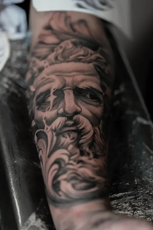 Tatuagem realista a preto e cinza do rosto de Zeus no antebraço