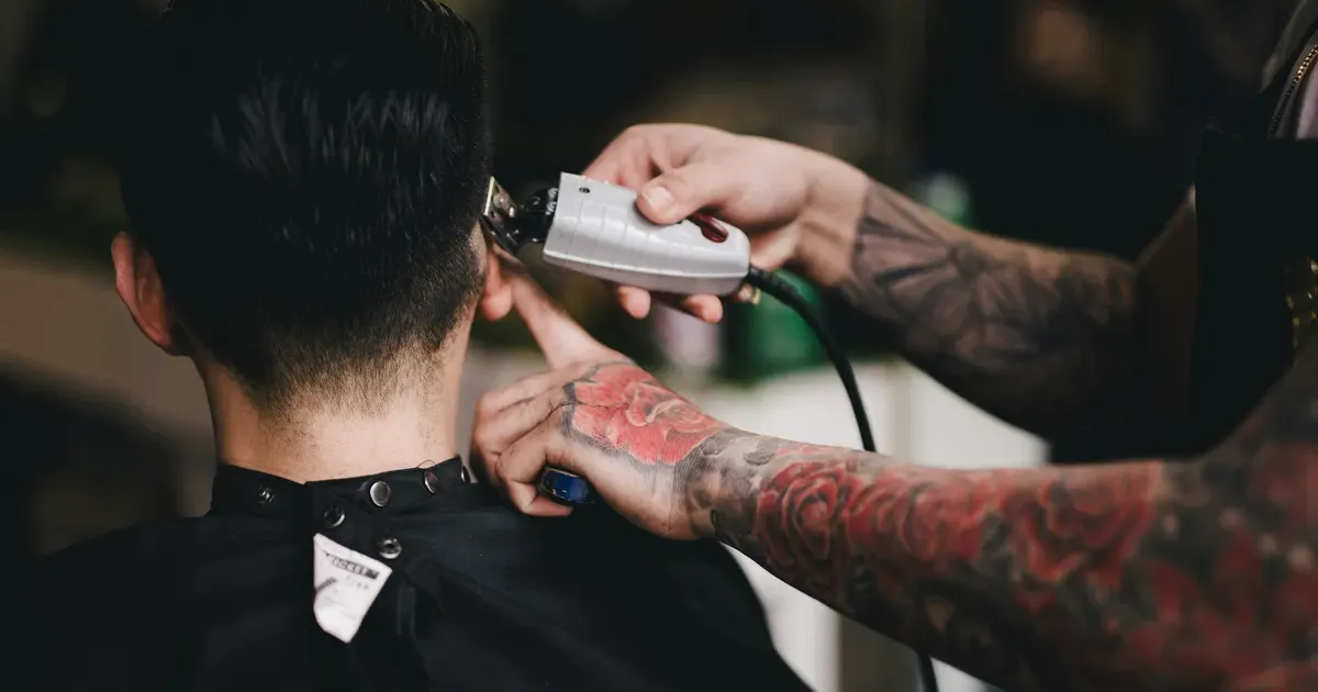 Barbeiro tatuado a fazer um fade com a máquina na nuca de um cliente